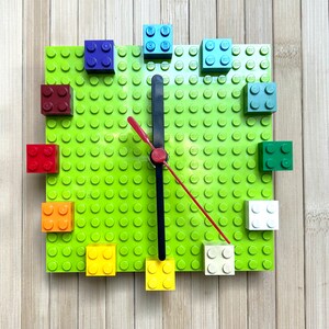 Może przedstawiać: Zielona płyta podstawowa Lego z tarczą zegara wykonaną z kolorowych klock&oacute;w Lego. Wskaz&oacute;wki zegara są czarne i czerwone.