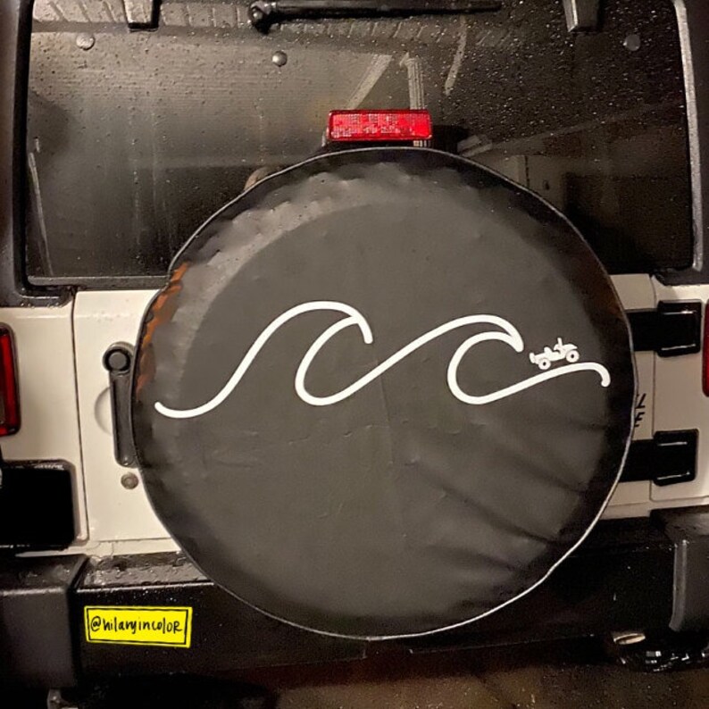 Puede incluir: Una cubierta de rueda de repuesto negra con un dise&ntilde;o de ola blanca y una peque&ntilde;a silueta de jeep blanca. La cubierta tiene el texto "@Hilaryincolor" en la parte inferior.