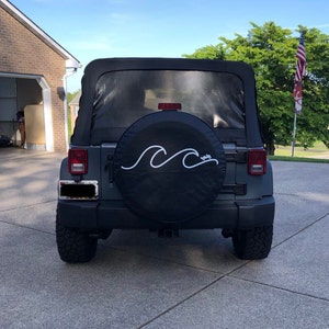 Puede incluir: Un Jeep Wrangler gris con una cubierta de rueda de repuesto negra que presenta un dise&ntilde;o de onda blanca. La cubierta tiene la palabra "salty" escrita en blanco en la esquina inferior derecha.