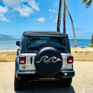 Puede incluir: Un Jeep Wrangler plateado con una rueda de repuesto negra que tiene un dise&ntilde;o de onda blanca. El Jeep est&aacute; estacionado en una playa de arena con palmeras en el fondo.