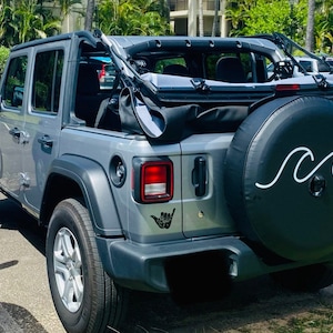 Puede incluir: Un Jeep Wrangler plateado con una cubierta de rueda de repuesto negra que presenta un dise&ntilde;o de ola blanca y el texto "Aloha". El Jeep tiene una capota blanda negra y un parachoques trasero negro. El Jeep est&aacute; estacionado en un estacionamiento.