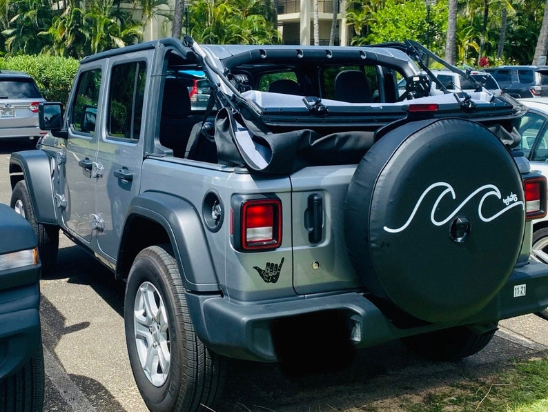 Puede incluir: Un Jeep Wrangler plateado con una cubierta de rueda de repuesto negra que presenta un dise&ntilde;o de ola blanca y el texto "Aloha". El Jeep tiene una capota blanda negra y un parachoques trasero negro. El Jeep est&aacute; estacionado en un estacionamiento.