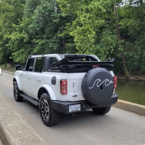 Puede incluir: Ford Bronco blanco con capota blanda negra y una cubierta de rueda de repuesto con un dise&ntilde;o de onda. La matr&iacute;cula dice "TXH-6668".