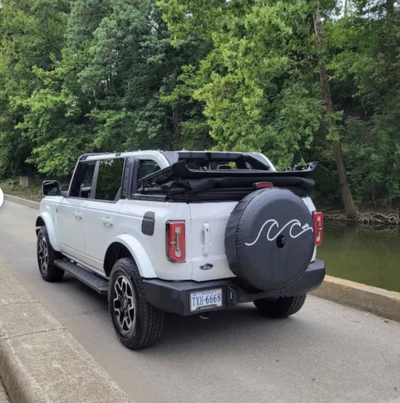 Puede incluir: Ford Bronco blanco con capota blanda negra y una cubierta de rueda de repuesto con un dise&ntilde;o de onda. La matr&iacute;cula dice "TXH-6668".