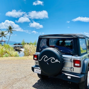 Puede incluir: Un Jeep plateado con una cubierta de rueda de repuesto negra que presenta un dise&ntilde;o de onda blanca. El Jeep est&aacute; estacionado en un camino de grava con vista al oc&eacute;ano y palmeras en el fondo.