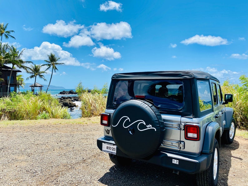 Puede incluir: Un Jeep plateado con una cubierta de rueda de repuesto negra que presenta un dise&ntilde;o de onda blanca. El Jeep est&aacute; estacionado en un camino de grava con vista al oc&eacute;ano y palmeras en el fondo.