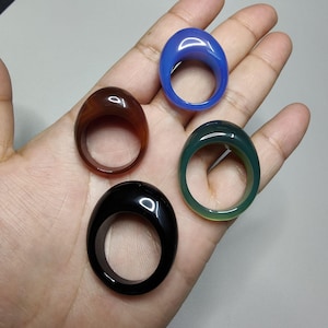 Puede incluir: Cuatro anillos ovalados de piedras preciosas. Los anillos están hechos de piedras preciosas azules, marrones, verdes y negras. Los anillos están en una mano.