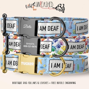 May include: Three dog collars with the text "I AM DEAF" on white labels. The collars have different patterns: blue with dog illustrations, colorful paw prints, and blue with bones. Buckles are engraved with names and phone numbers.