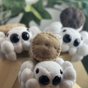 May include: Close-up of several plush spider toys with white bodies, large black eyes, and brown accents. The spiders have multiple legs and are arranged on a light brown surface. The toys are soft and cuddly.
