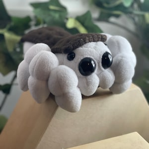 May include: White plush toy with brown felt on top, resembling a creature with large black eyes and a rounded body.