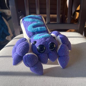 May include: A plush toy spider with a white body, purple legs, and blue accents. The spider has large black eyes and a friendly expression.