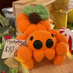 May include: Orange plush toy shaped like a spider with a pumpkin on its head. The pumpkin has a green leaf on top. The toy has large black eyes and a price tag that says "Pumpkin $80".