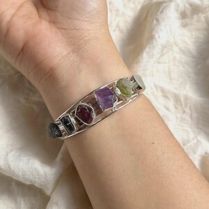 May include: A silver-toned cuff bracelet featuring a series of five raw, uncut gemstones in varying colors including dark gray, deep red, purple, light green, and a pale translucent stone, all set within a simple open-ended metal frame.