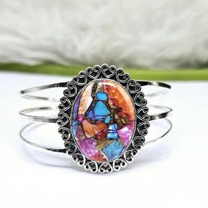 Peut inclure: Un bracelet manchette en argent avec une grande pierre ovale sertie dans un cadre décoratif en forme de cœur. La pierre est un mélange de turquoise, rose, orange et marron.