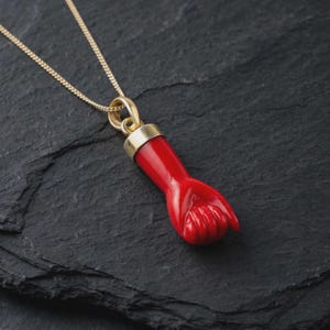 Puede incluir: Un collar de cadena dorada con un colgante en forma de puño cerrado. El puño es de un color rojo vibrante y está unido a la cadena dorada mediante una anilla de metal dorado pulido. El colgante está colocado en ángulo, mostrando los dedos.