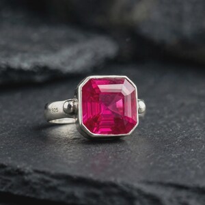 Puede incluir: Un anillo de plata de ley con una gran piedra preciosa de color rojo rosado intenso, de corte cuadrado y engastada en un bisel. La banda de plata pulida presenta un sello 925 claramente visible en el lateral, indicando la pureza del metal.