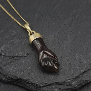 May include: A dark brown, carved stone pendant shaped like a clenched fist, suspended from a thin gold-toned chain. The pendant features a smooth, polished finish and is attached to the chain by a simple, cylindrical gold-toned metal bail.