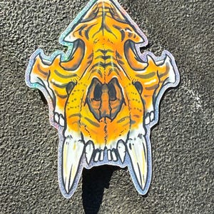 May include: A sticker featuring a stylized tiger skull design. The skull is primarily orange with black stripes and details, and white fangs. The sticker has a holographic, iridescent border, giving it a shimmering effect.