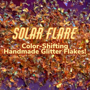 May include: Close-up of iridescent, color-shifting glitter flakes. The glitter is a mix of gold, orange, and red hues. The text "SOLAR FLARE Color-Shifting Handmade Glitter Flakes!" is superimposed on the image.