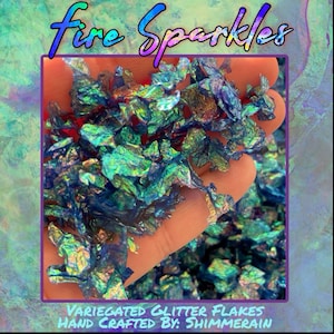 May include: A close-up of iridescent teal, green, and blue glitter flakes. The glitter is in a hand, and the text "Fire Sparkles" is at the top of the image. The text "VARIEGATED GLITTER FLAKES HAND CRAFTED BY: SHIMMERAIN" is at the bottom of the image.