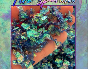 May include: A close-up of iridescent teal, green, and blue glitter flakes. The glitter is in a hand, and the text "Fire Sparkles" is at the top of the image. The text "VARIEGATED GLITTER FLAKES HAND CRAFTED BY: SHIMMERAIN" is at the bottom of the image.