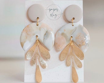 May include: A pair of gold and white statement earrings with a marbled design and gold leaf accents. The earrings are displayed on a white card with the text "paper clay".