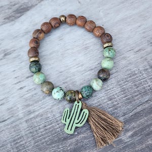 May include: A green cactus charm with a brown tassel hangs from a bracelet made of brown wood beads and green turquoise beads.