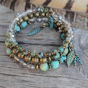 Könnte beinhalten: Ein Stapel von drei Armbändern mit Perlen. Die Armbänder haben türkisfarbene, klare und goldfarbene Perlen. Dekorative Blattanhänger hängen an den Armbändern. Die Armbänder werden auf einer Holzoberfläche präsentiert.