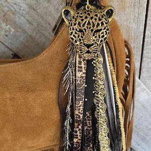 May include: A tan suede fringe bag featuring a decorative gold-tone leopard head charm attached to a metal lobster clasp. The charm is adorned with sparkling crystals and hangs above a cluster of layered ribbons in black, gold, and leopard print.