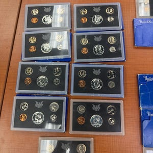 May include: A collection of United States proof sets in clear plastic cases, each containing various coins. The sets include silver and copper-colored coins, with a blue box in the background. The coins feature portraits and emblems.