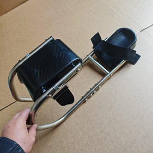 May include: A silver-colored mobility device with black padded supports and straps. The device has a curved frame and is designed to provide support and stability. The image shows the device on a brown surface.