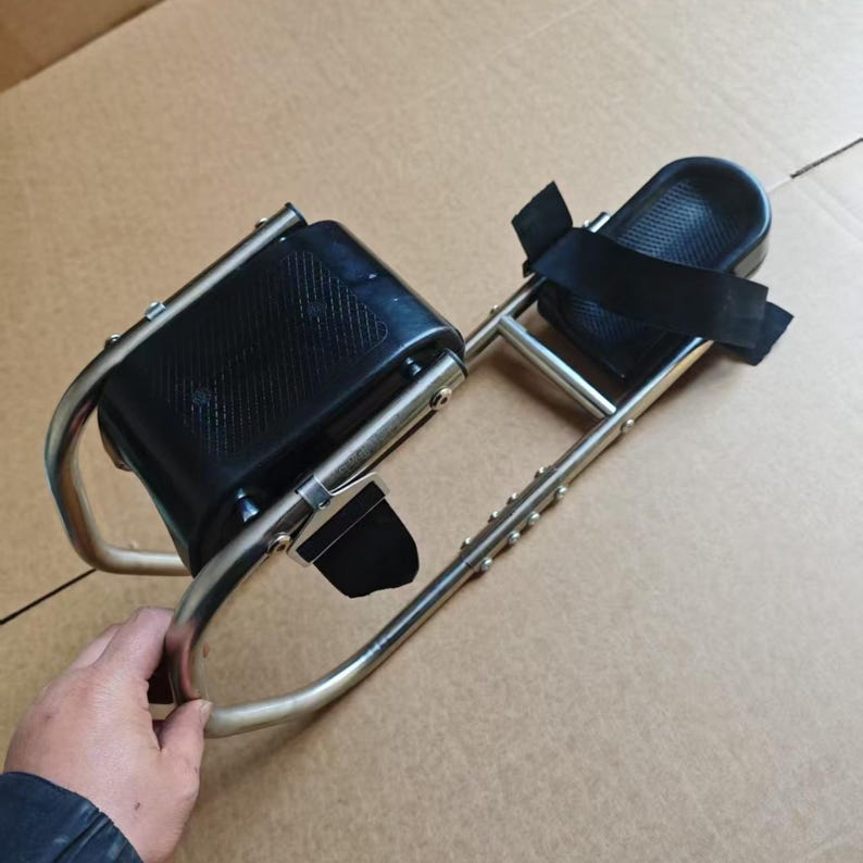 May include: A silver-colored mobility device with black padded supports and straps. The device has a curved frame and is designed to provide support and stability. The image shows the device on a brown surface.