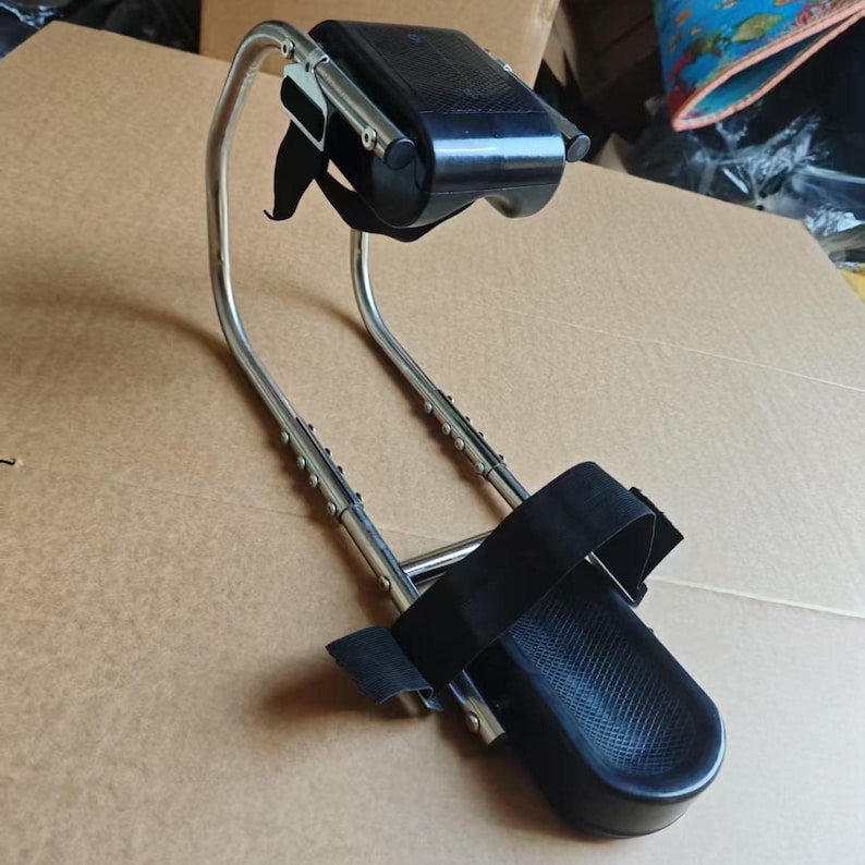May include: A silver-colored metal medical device with black padded supports for the foot and leg. The device has adjustable straps and a curved frame, designed for patient support and mobility.