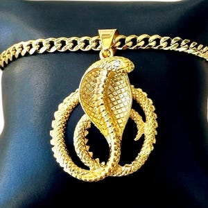Peut inclure: Un collier pendentif doré avec un motif de cobra. Le cobra est enroulé et présente un motif d'écailles détaillé. Le collier a une chaîne gourmette et est présenté sur une surface noire.