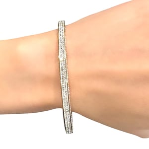 May include: A silver-toned bangle bracelet with a row of small, clear gemstones. The bracelet is a simple, elegant design, perfect for everyday wear. The bracelet is shown on a person's arm.