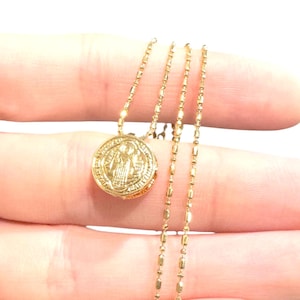 Puede incluir: Un collar dorado con un colgante redondo con un diseño en relieve y una cadena delicada. El colgante es el punto focal, mostrando detalles intrincados. La cadena es fina y elegante, lo que aumenta el atractivo general del collar.