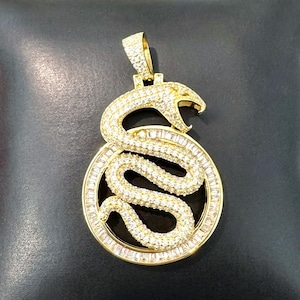 May include: A gold pendant with a snake design, set with white gemstones. The snake is coiled inside a circular frame, also with gemstones. The pendant is displayed on a black surface.