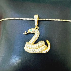 Peut inclure: Un collier pendentif serpent doré avec un motif de serpent enroulé recouvert de petites pierres pavées transparentes. Le serpent possède une petite pierre verte en guise d'œil. Le pendentif est suspendu à une fine chaîne en boîte dorée.