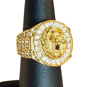 Op de afbeelding: Een goudkleurige ring met een cirkelvormig voorvlak met in het midden een sculpturaal reliëf van een leeuwenkop, geaccentueerd door een kleine rode steen. De leeuw is omringd door een rand van heldere, rondgeslepen stenen en de band is bezet met rijen heldere stenen.