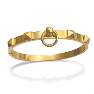 May include: A gold-toned stainless steel hinged bangle bracelet featuring a central circular ring element and four evenly spaced pyramid-shaped studs along the band. The metal surface is polished and reflective, with the word Stainless engraved.