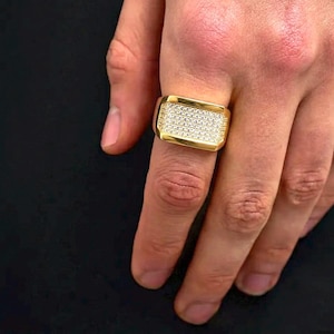 May include: A gold-toned rectangular ring featuring a flat top surface completely encrusted with numerous small, clear, sparkling stones arranged in a dense grid pattern, set within a polished metal band that wraps around the finger.