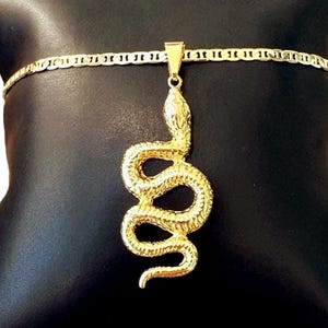 Op de afbeelding: Een goudkleurige slangenhangerketting. De hanger is een gedetailleerd, opgerold slangenontwerp. De ketting is een platte, geschakelde stijl. De sieraden worden gepresenteerd op een zwart kussen.