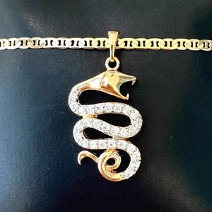 Op de afbeelding: Goudkleurige armband met een slangenhanger. De slangenhanger is versierd met heldere kristallen en heeft een gedetailleerde kop. De armband wordt gepresenteerd op een zwarte ondergrond.