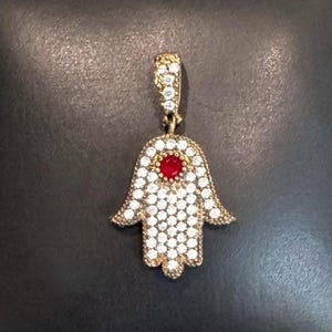 May include: A gold-toned Hamsa hand pendant featuring a central round red stone surrounded by numerous small, clear, sparkling pavé-set stones. The pendant includes a matching gold-toned bail at the top, also encrusted with small clear stones.