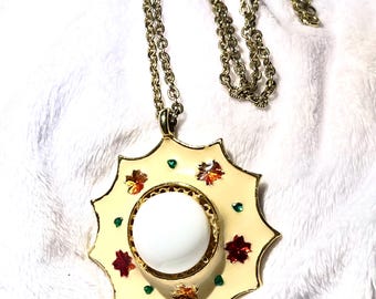 May include: A gold-tone chain necklace featuring a star-shaped pendant with a cream-colored enamel finish. The pendant is centered with a smooth white cabochon and decorated with small, colorful leaf-shaped accents and tiny green dots.