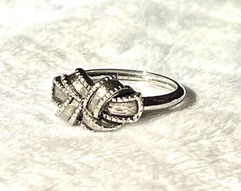 May include: A silver-toned ring featuring a central knot design with a textured, brushed metal finish. The knot element is detailed with small, raised beaded edges along its loops, creating a dimensional appearance on the thin, polished band.