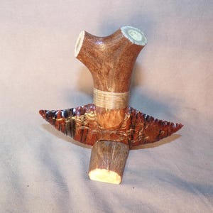 May include: A decorative object featuring a dark, reddish-brown, serrated stone blade attached to a Y-shaped antler handle. The handle is wrapped with light-colored cordage at the center, and the base of the antler rests on a flat surface.
