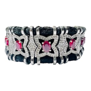 May include: A wide cuff bracelet featuring a black textured band accented with silver-toned metal links. Each link displays a four-pointed star shape encrusted with small clear stones and a central oval-cut red gemstone set in the middle.