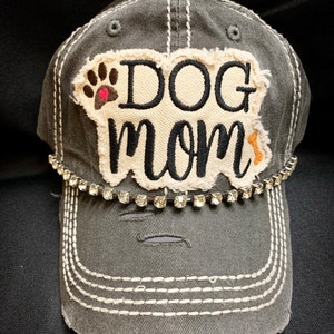 K&ouml;nnte beinhalten: Eine graue, abgenutzte Baseballkappe mit einem wei&szlig;en Patch, auf dem "DOG mom" in schwarzer Stickerei steht. Der Patch hat einen braunen Pfotenabdruck mit einem rosa Herzen und einem kleinen orangenen Knochen. Die Kappe hat eine Reihe von klaren Strasssteinen um den Rand.