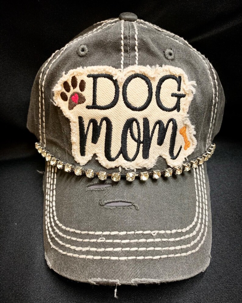 K&ouml;nnte beinhalten: Eine graue, abgenutzte Baseballkappe mit einem wei&szlig;en Patch, auf dem "DOG mom" in schwarzer Stickerei steht. Der Patch hat einen braunen Pfotenabdruck mit einem rosa Herzen und einem kleinen orangenen Knochen. Die Kappe hat eine Reihe von klaren Strasssteinen um den Rand.